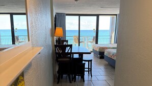 A room with a dining table and chairs, a bed with a colorful bedspread, and a view of the ocean through large windows. - Spacious Ocean Front Studio for 4 with Picture-Perfect View! (Daytona Beach Shores)