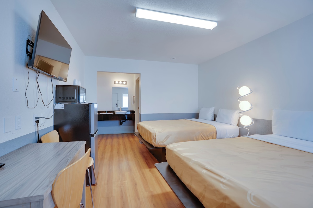 A hotel room with a desk, two beds, a television, and a bathroom with a mirror and lights.