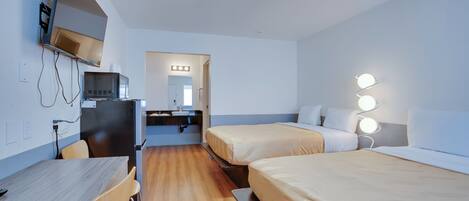 A hotel room with a desk, two beds, a television, and a bathroom with a mirror and lights.