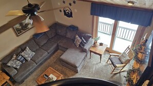 Un salon chaleureux avec un canapé d’angle, une petite table, un fauteuil à bascule et un ventilateur de plafond.