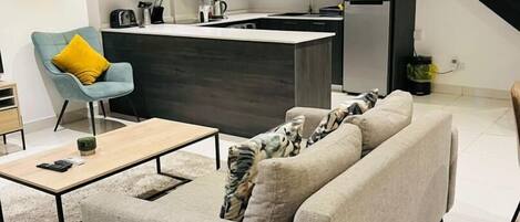 Un salon moderne avec un canapé gris, une table basse en bois et un fauteuil bleu agrémenté d’un coussin jaune. On aperçoit, au fond, une cuisine avec des meubles sombres et un escalier noir.