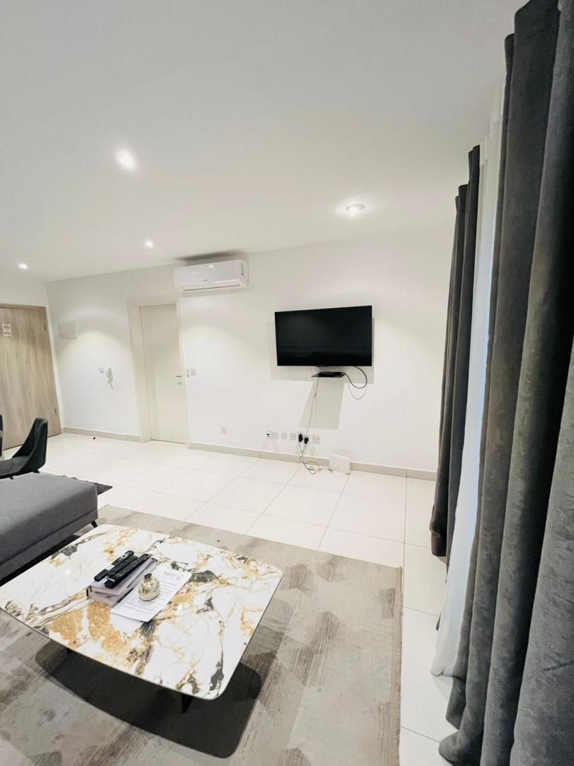 En moderne stue med sofa, sofabord, fladskærms-tv monteret på væggen og et stort vindue med gardiner.