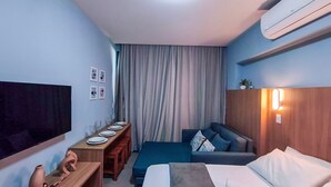 A hotel room with a bed, a sofa, a desk with a flat-screen TV, a wooden side table, and a large window with curtains. - Flats Garden no Nomar Carneiros Ecoloving (Tamandaré)