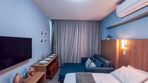 A hotel room with a bed, a sofa, a desk with a flat-screen TV, a wooden side table, and a large window with curtains. - Flats Garden no Nomar Carneiros Ecoloving (Tamandaré)