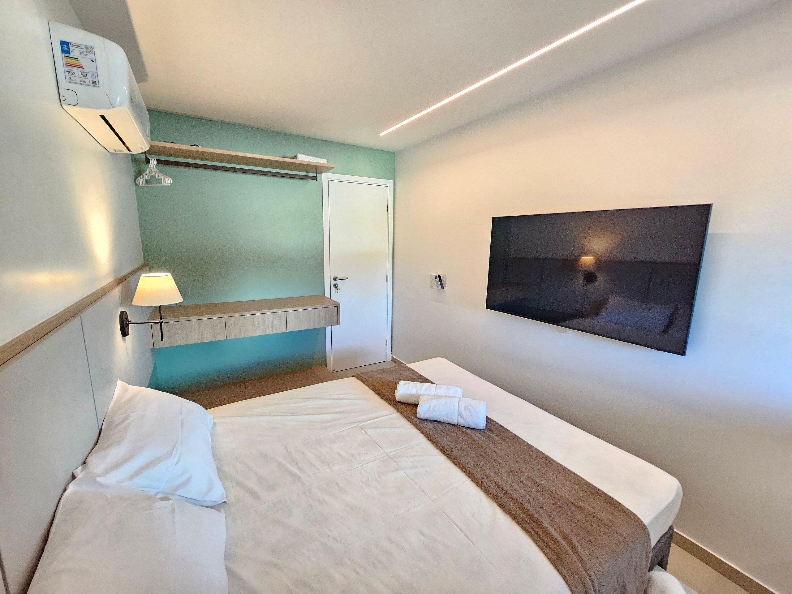 A hotel room with a bed, a mounted TV, a wall-mounted air conditioner, and a towel on the bed.