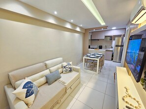 A modern living room with a sofa, a dining table, and a kitchen area in the background. - Galapagos Beach Residence - Praia dos Carneiros (Tamandaré)