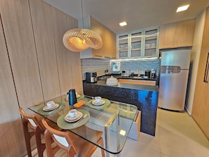 A modern kitchen with a glass dining table, wooden chairs, a coffee maker, and a stainless steel refrigerator.