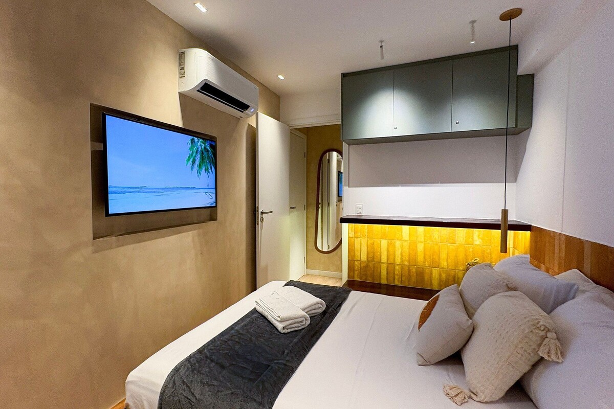 A hotel room with a bed, a television mounted on the wall, and an air conditioning unit.
