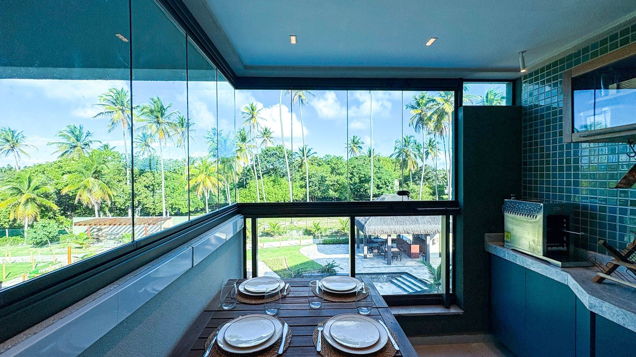 A balcony with a table set for dining, overlooking a tropical landscape with palm trees and a thatched-roof structure.