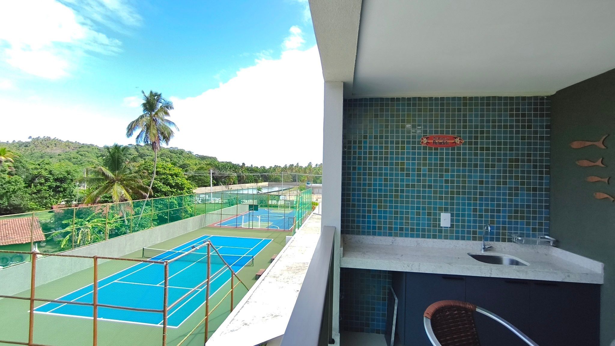 Un balcón con cancha de tenis, vistas a un paisaje tropical y una pared de azulejos con un letrero.