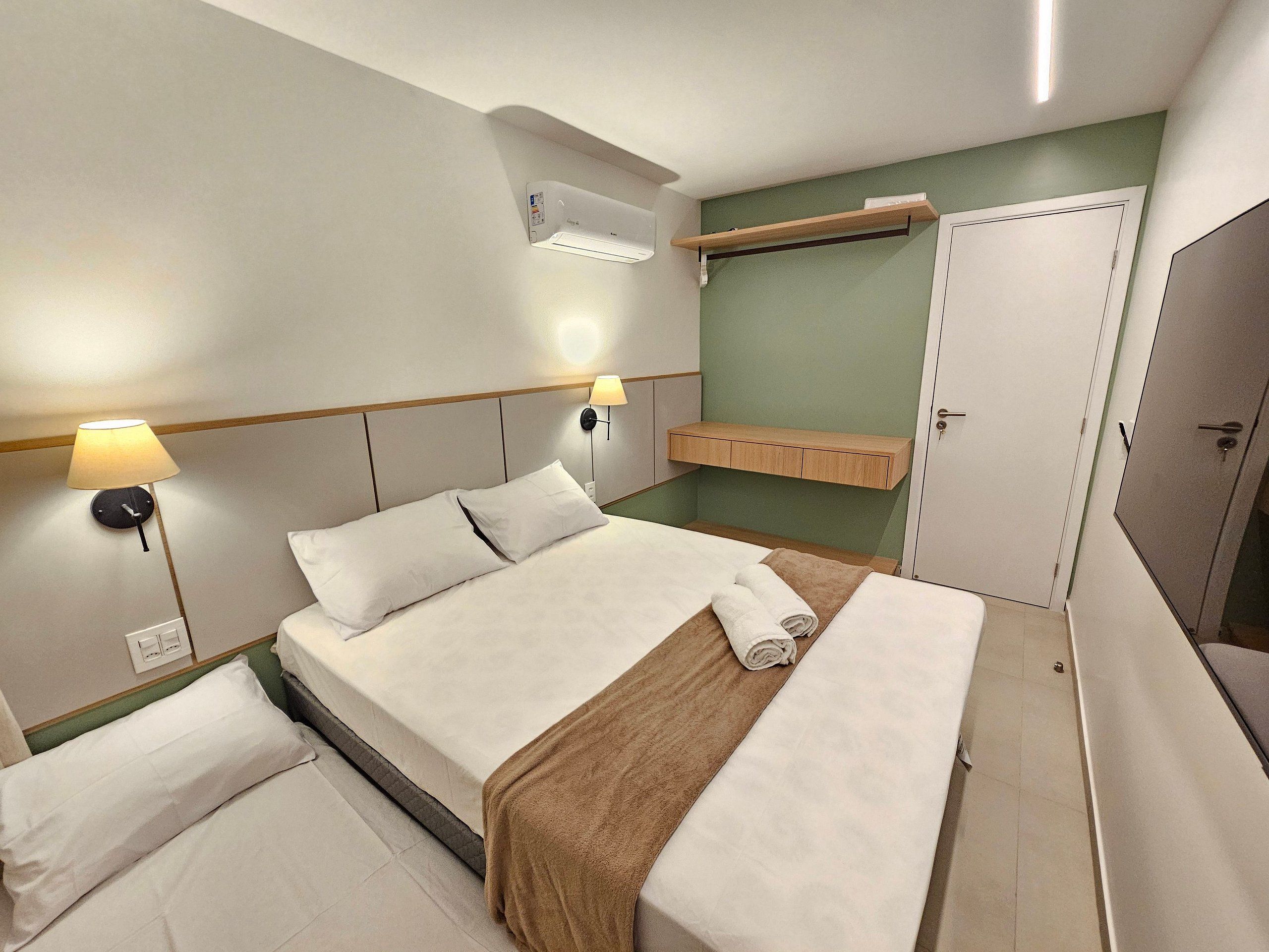 A hotel room with a bed, bedside lamps, an air conditioning unit, a shelf, and a wardrobe.