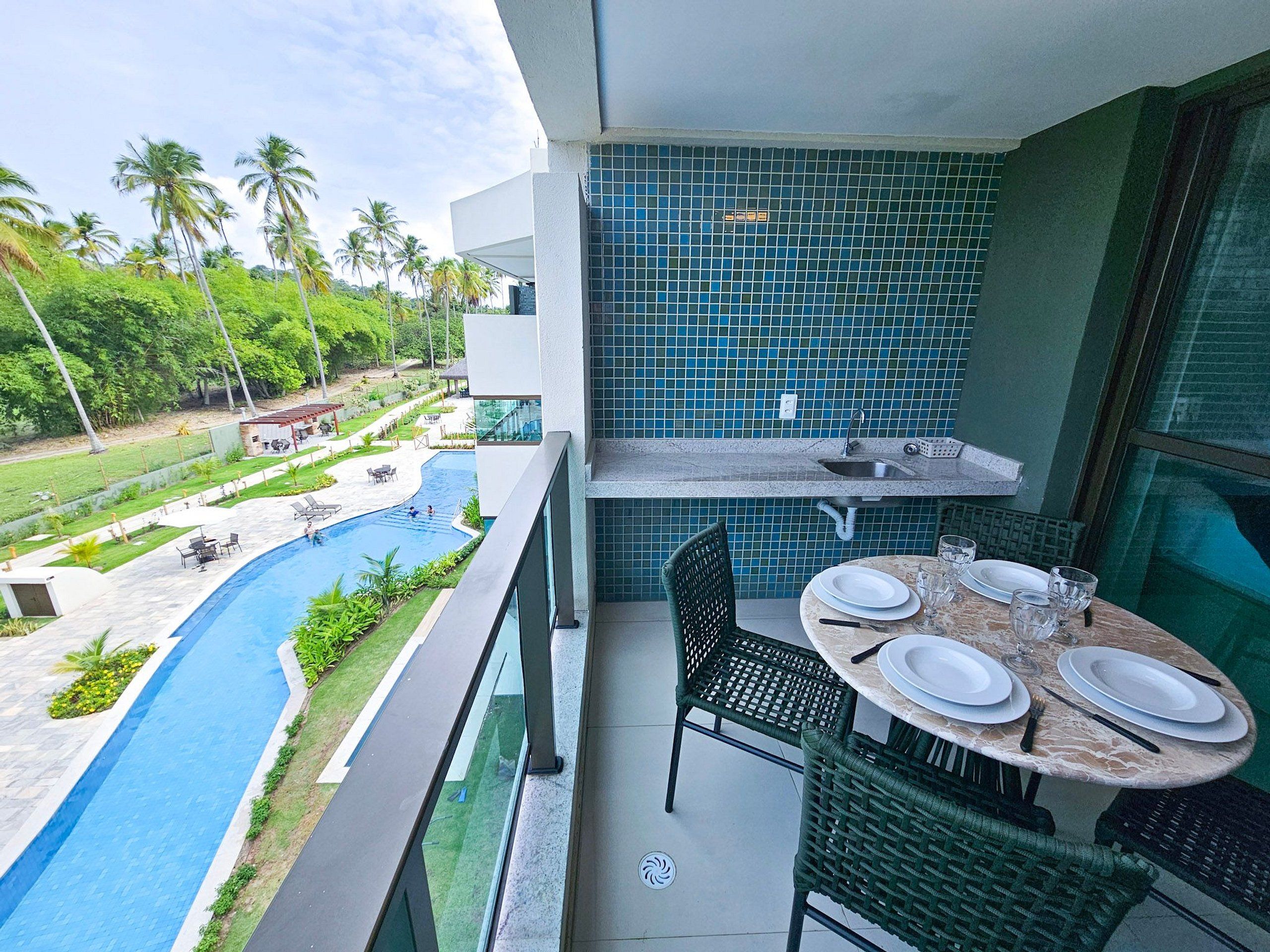 A balcony with a view of a swimming pool, a dining table set with plates and glasses, and a tiled backsplash.