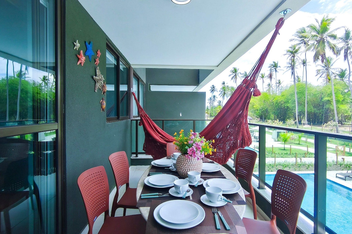 A dining area with a table set for four, chairs, a pool, and a view of palm trees.