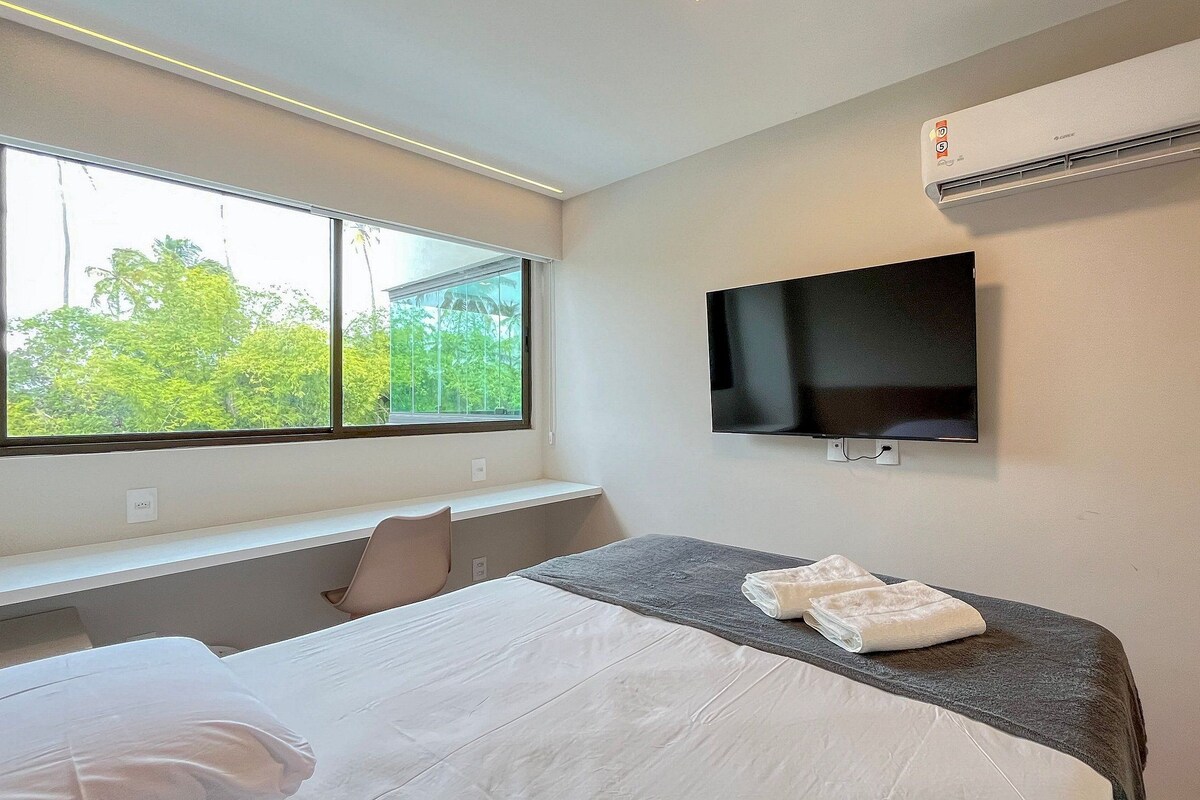 A hotel room with a bed, a flat-screen TV, an air conditioner, and a window with a view of greenery.