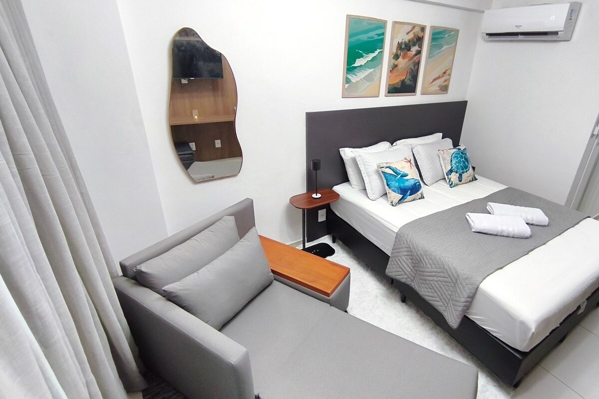 A hotel room with a bed, a chair, a nightstand, a mirror, and artwork on the wall.