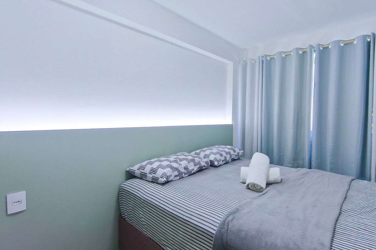 A single bed with striped bedding, a pillow, and a blanket. There is a power outlet on the wall.