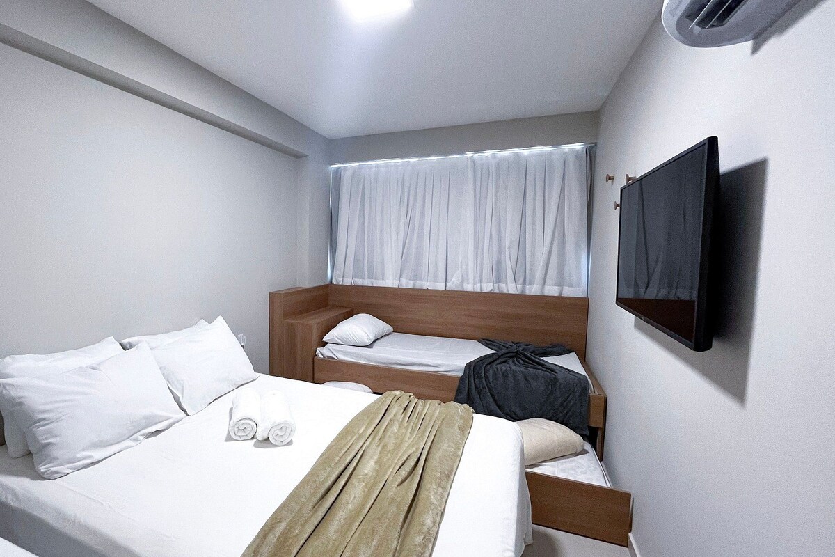A small, single-bed room with a wooden headboard, a flat-screen TV mounted on the wall, and a window with white curtains.
