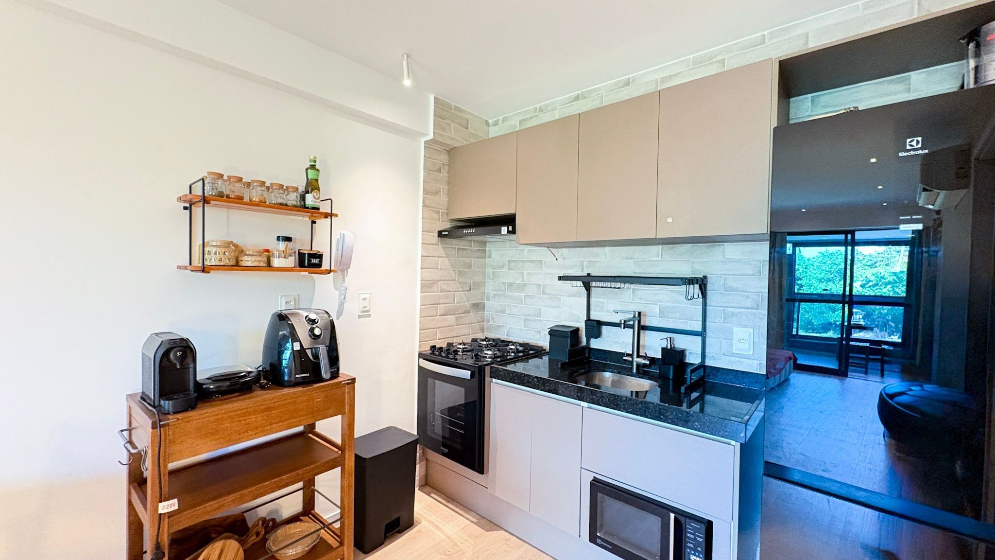 A compact kitchen with a stove, microwave, and a coffee machine. There is a wooden shelf with jars and a phone. The kitchen has a modern design with a brick backsplash and a large window.