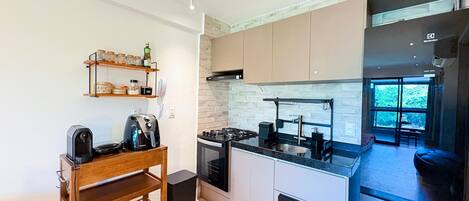 A compact kitchen with a stove, microwave, and a coffee machine. There is a wooden shelf with jars and a phone. The kitchen has a modern design with a brick backsplash and a large window.