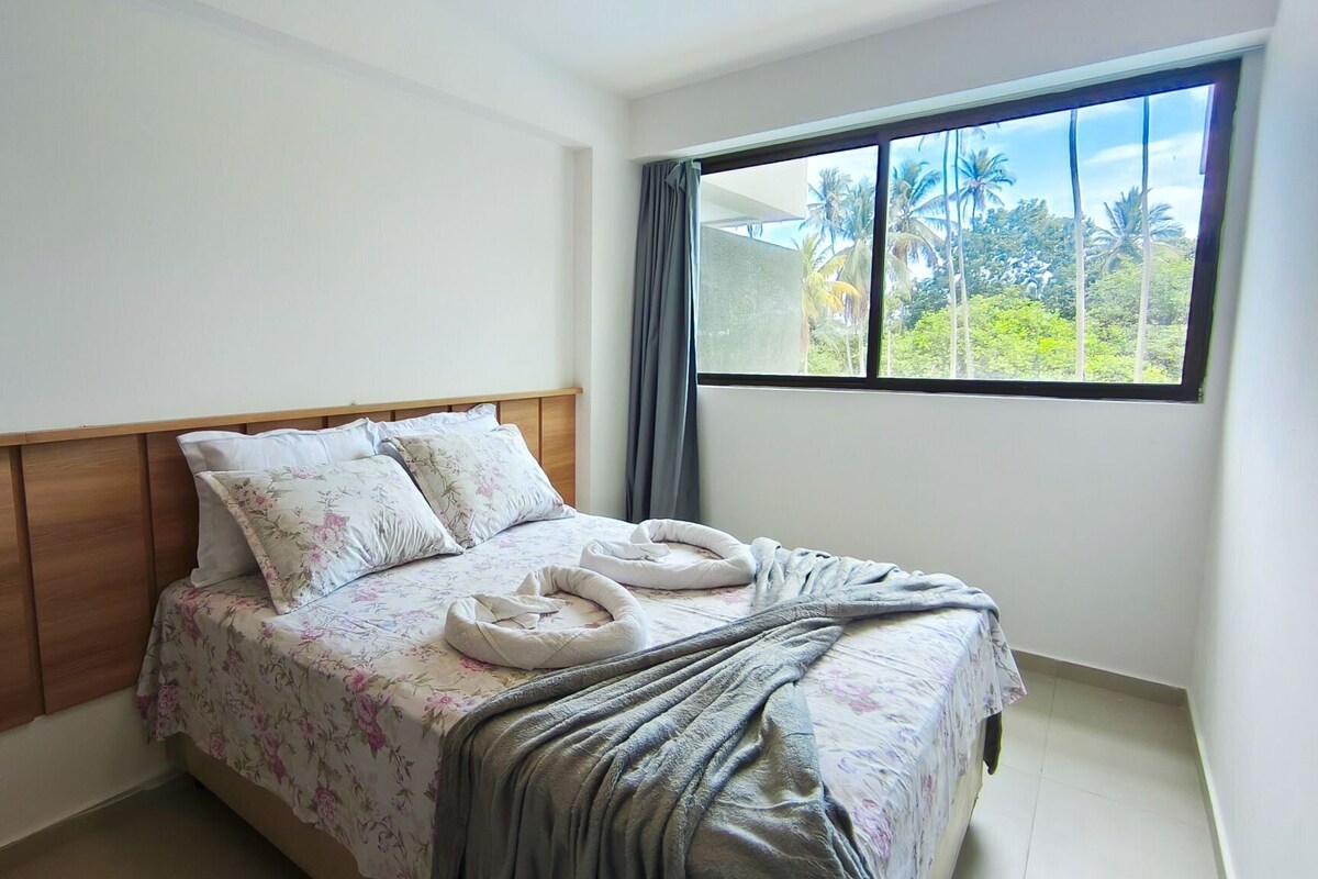 A bedroom with a wooden headboard, floral bedding, a window with a view of palm trees, and a towel on the bed.