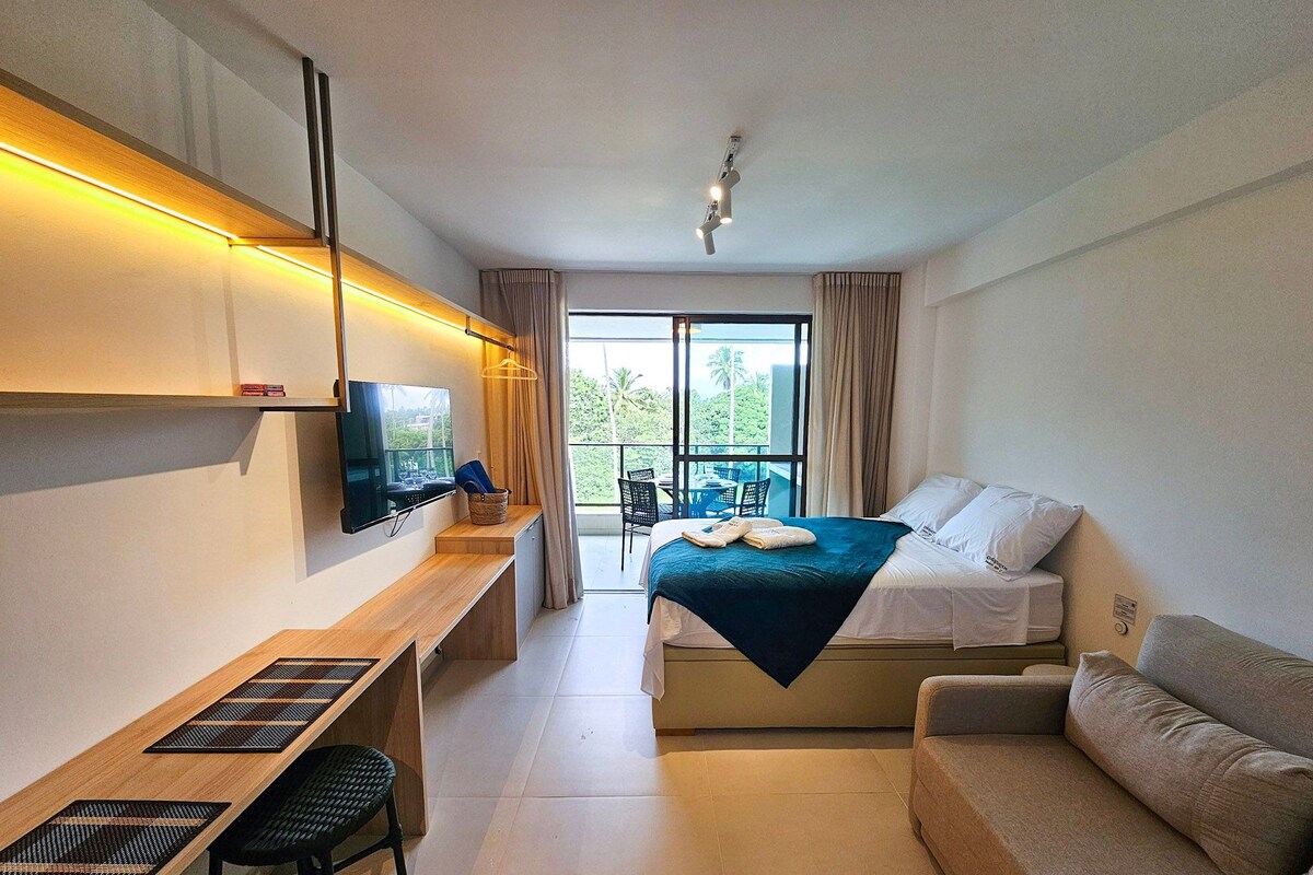 A hotel room with a bed, a desk with a TV, a sofa, and a balcony with a view.