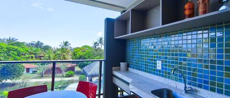 A balcony with a table and chairs, a kitchen area with blue tiles, and a view of greenery and buildings.
