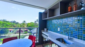 A balcony with a table and chairs, a kitchen area with blue tiles, and a view of greenery and buildings. - Galapagos Beach Residence - Praia dos Carneiros (Tamandaré)