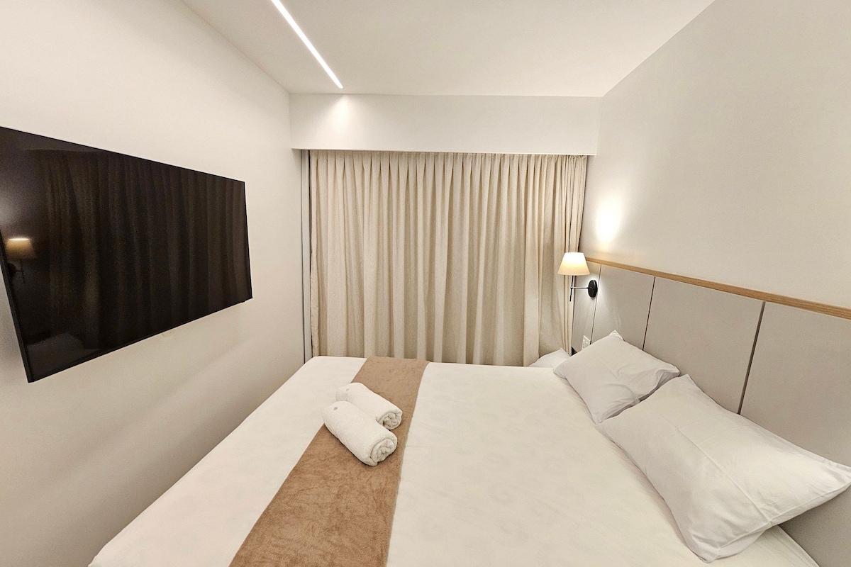 Ein Hotelzimmer mit Bett, Wandfernseher, Nachttischlampe und einem fenster mit Vorhängen.