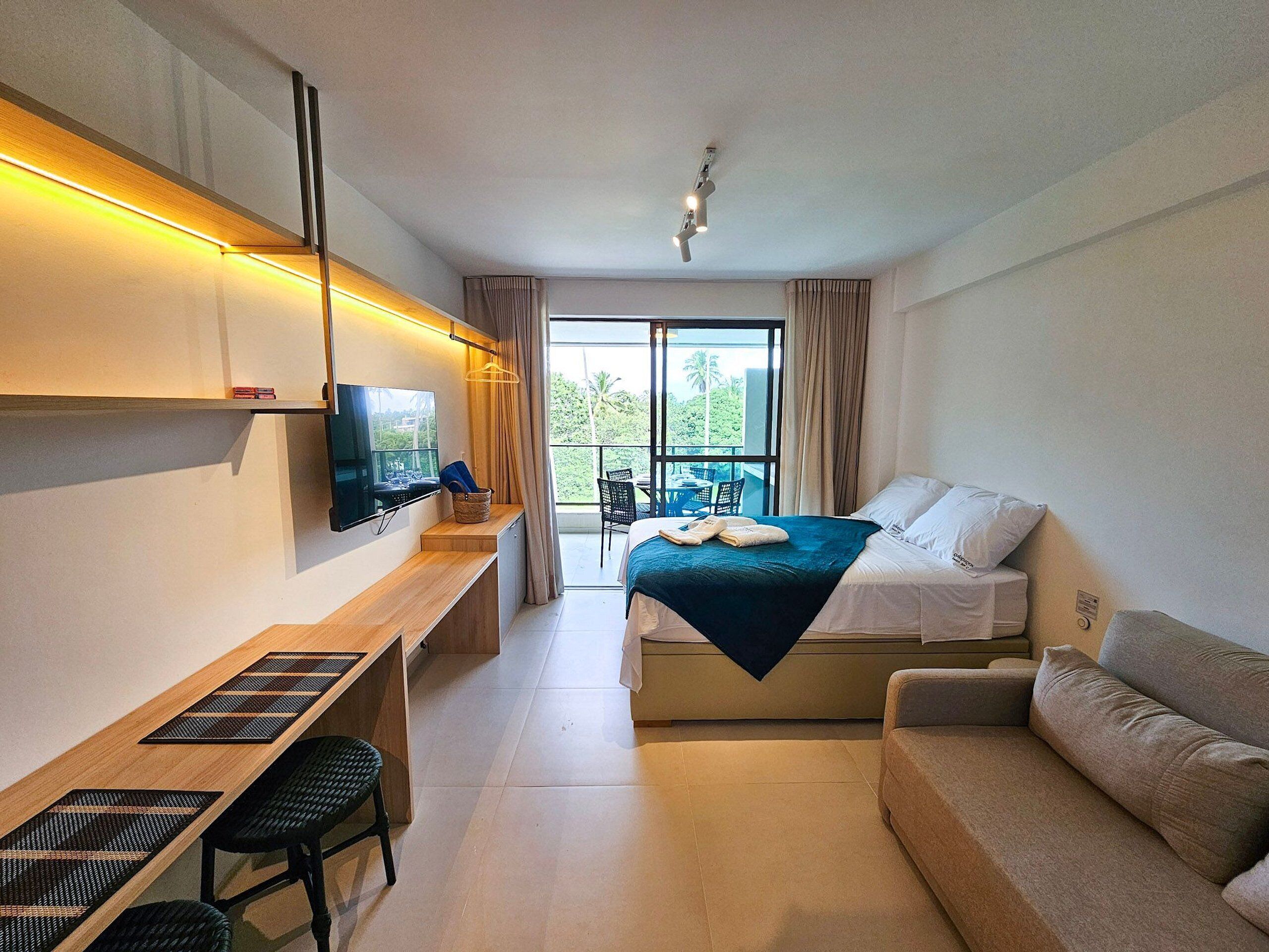 A hotel room with a bed, a desk with a TV, a sofa, and a balcony with a view.