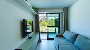A modern living room with a flat-screen TV, a grey sofa, a sliding glass door leading to a balcony, and a small plant on a shelf. - Galapagos Beach Residence - Praia dos Carneiros (Tamandaré)