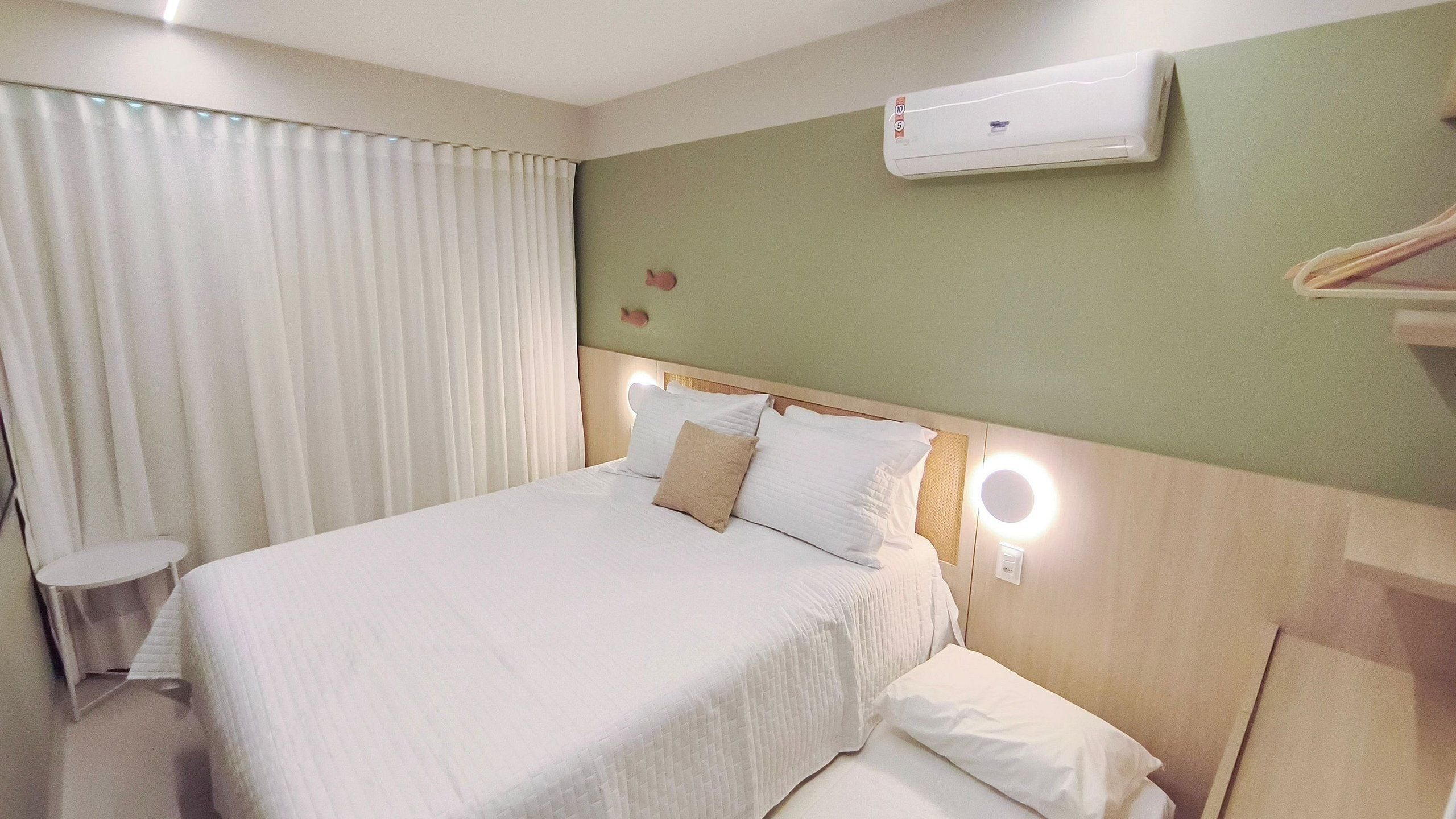 A hotel room with a bed, a bedside table, a wall-mounted air conditioner, and a window with curtains.