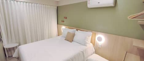 A hotel room with a bed, a bedside table, a wall-mounted air conditioner, and a window with curtains.