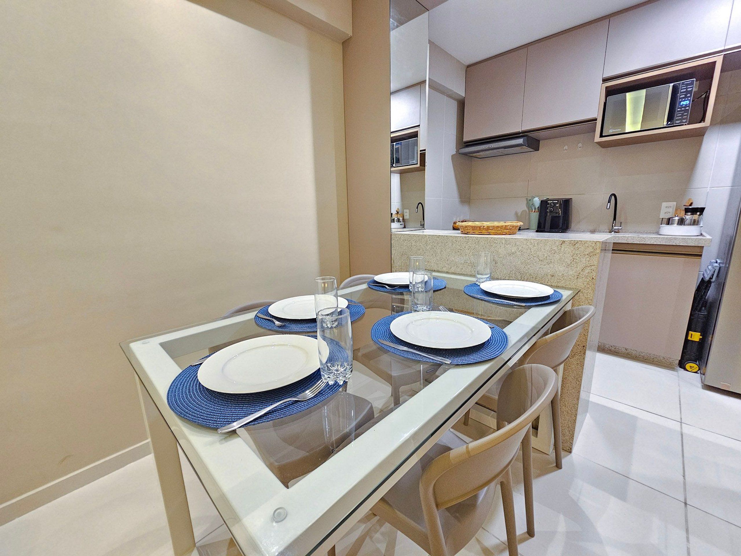 Uma sala de jantar com mesa de vidro, jogos americanos azuis, pratos brancos e copos transparentes.
