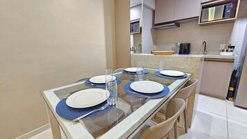 Uma sala de jantar com mesa de vidro, jogos americanos azuis, pratos brancos e copos transparentes.