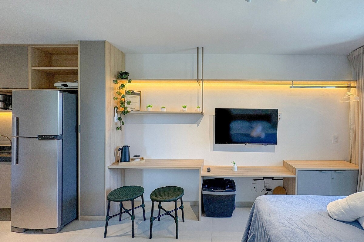 Habitación de hotel compacta con kitchenette, una cama, un escritorio con dos sillas, un televisor y una ventana con cortinas.