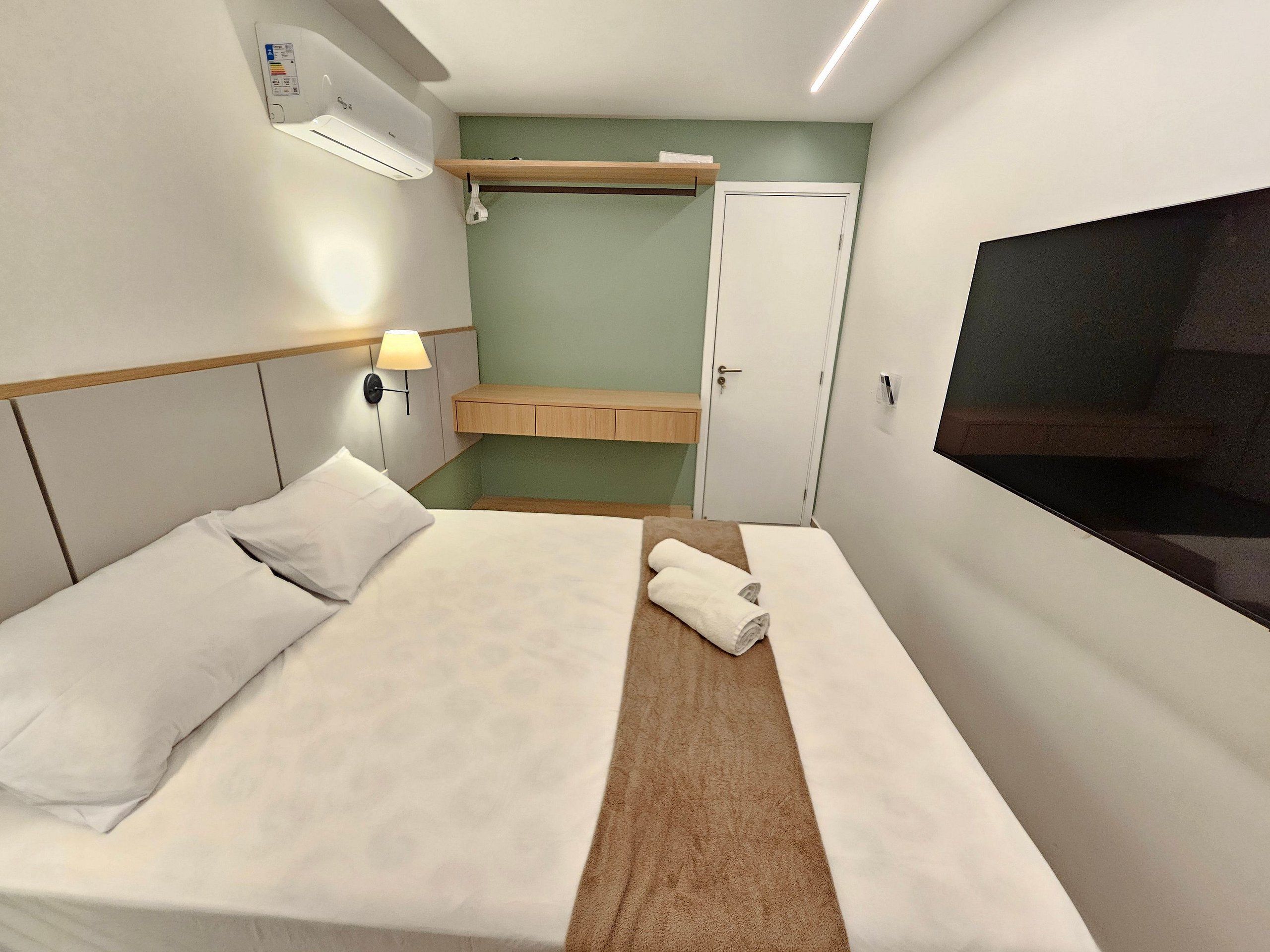 A hotel room with a bed, a wall-mounted TV, a small shelf, a door, and an air conditioning unit.