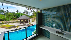 A balcony with a pool, a kitchen area with blue tiles, and a view of a resort with a thatched-roof structure. - Galapagos Beach Residence - Praia dos Carneiros (Tamandaré)