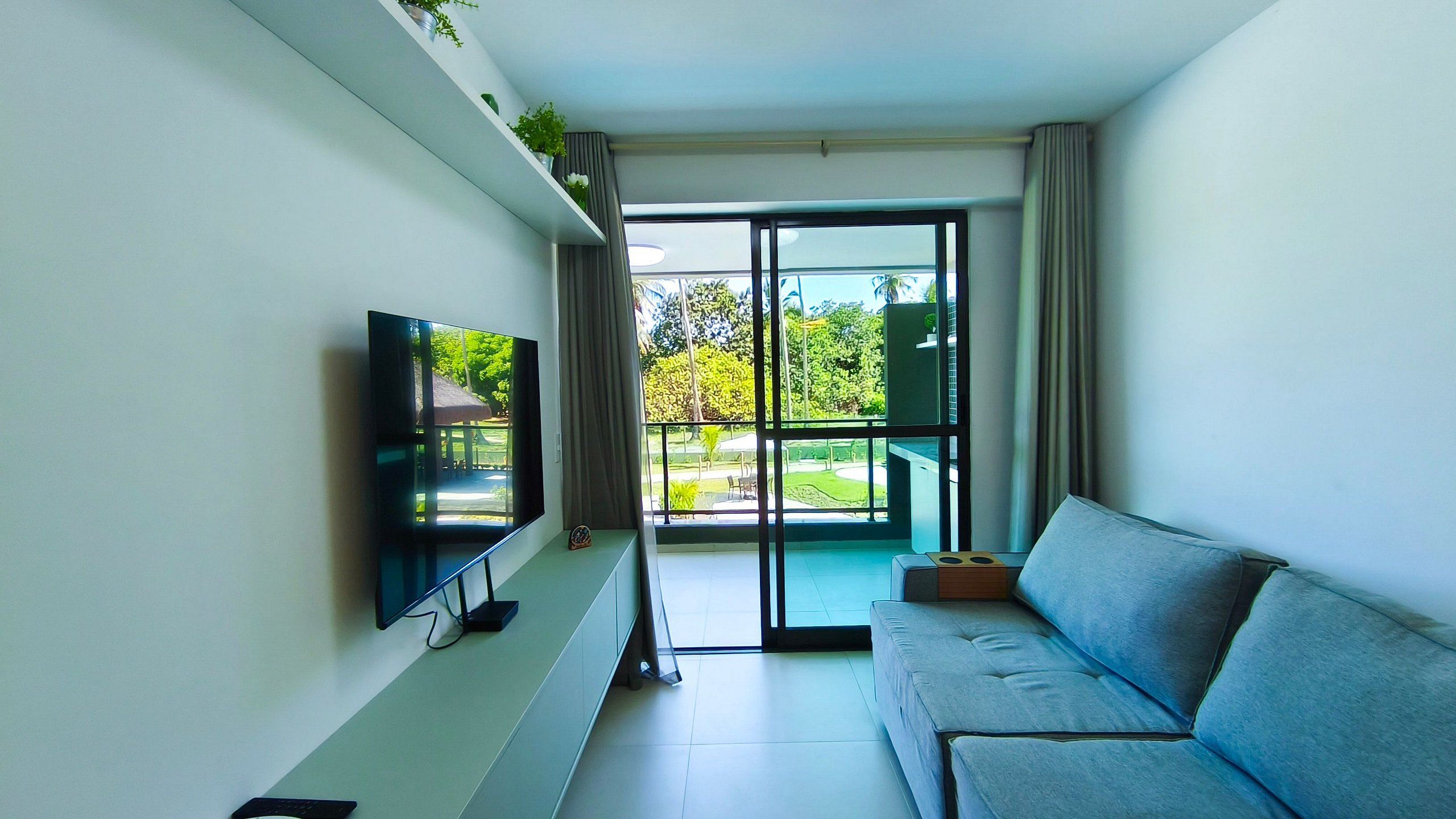 A modern living room with a flat-screen TV, a grey sofa, a sliding glass door leading to a balcony, and a small plant on a shelf.