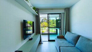 A modern living room with a flat-screen TV, a grey sofa, a sliding glass door leading to a balcony, and a small plant on a shelf.