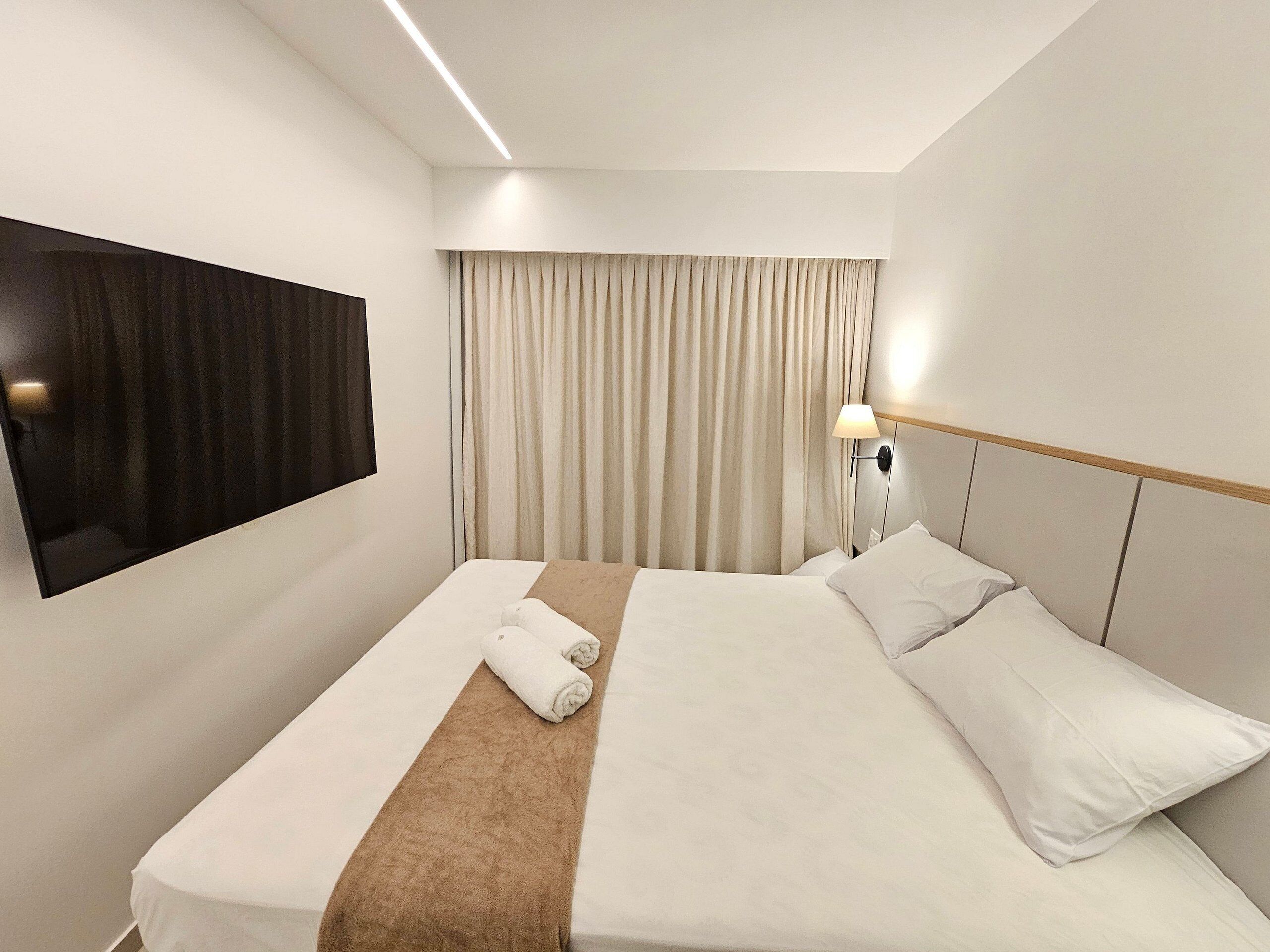 A hotel room with a bed, a TV mounted on the wall, a bedside lamp, and a window with curtains.