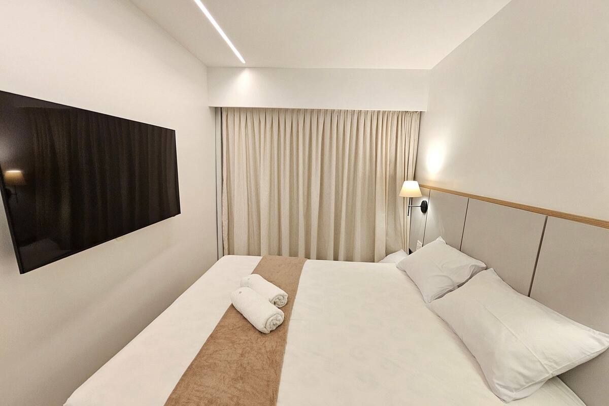 Una habitación de hotel con cama, televisión montada en la pared, lámpara de noche y ventana con cortinas.