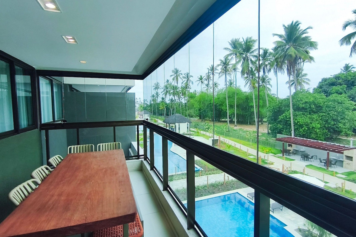A balcony with a wooden table and chairs, overlooking a swimming pool and lush greenery.