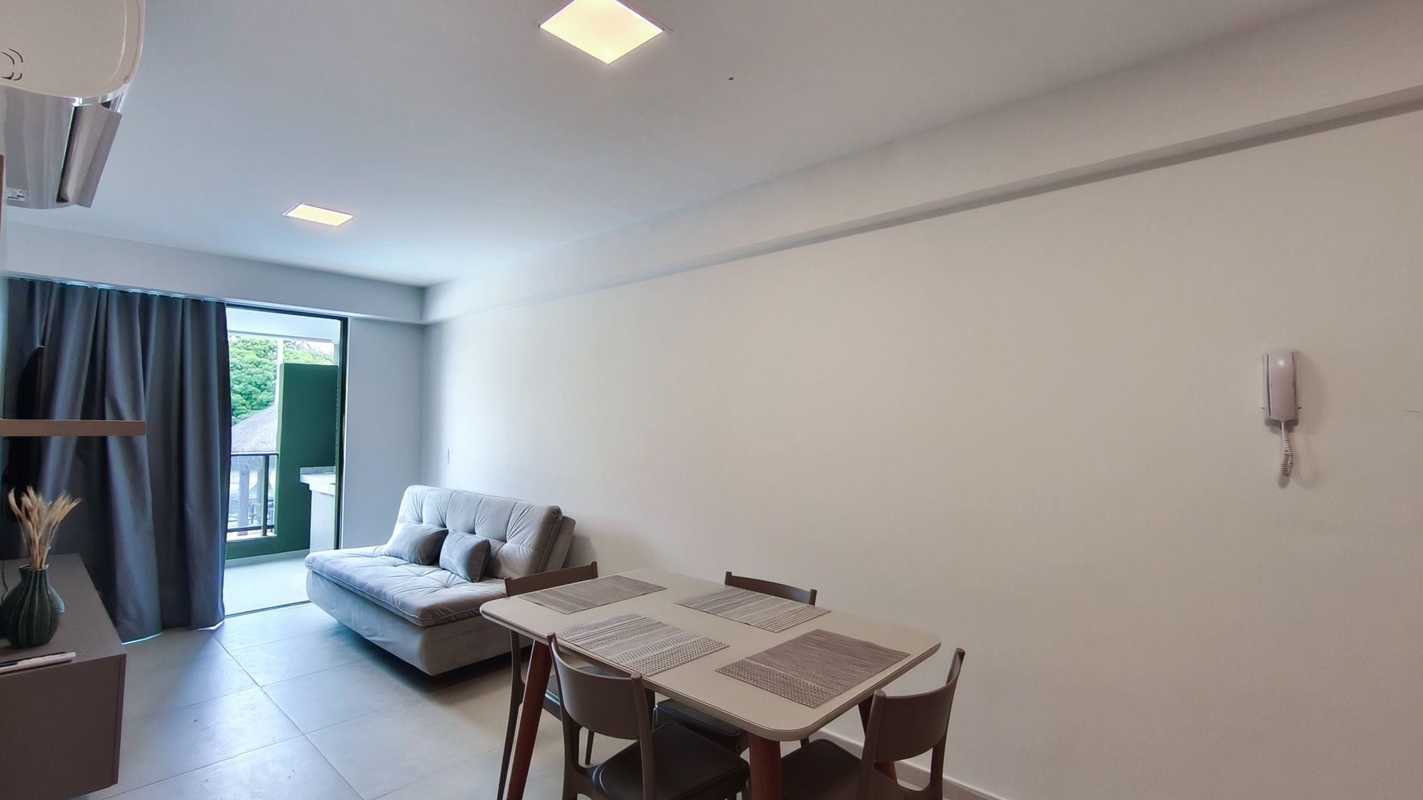A modern living room with a sofa, a dining table with chairs, a small kitchen area, and a sliding glass door leading to an outdoor space.