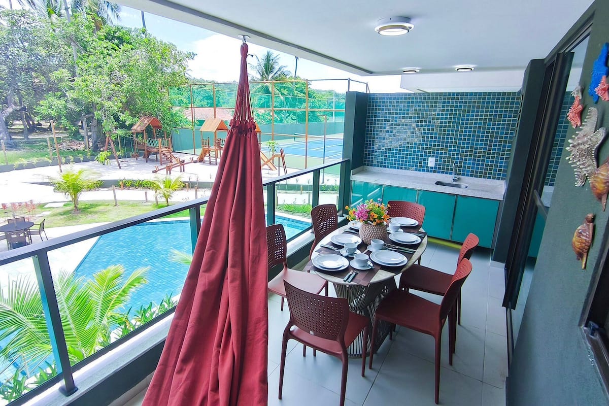 Balcón con una mesa de comedor para cuatro, con vista a una piscina y abundante vegetación.