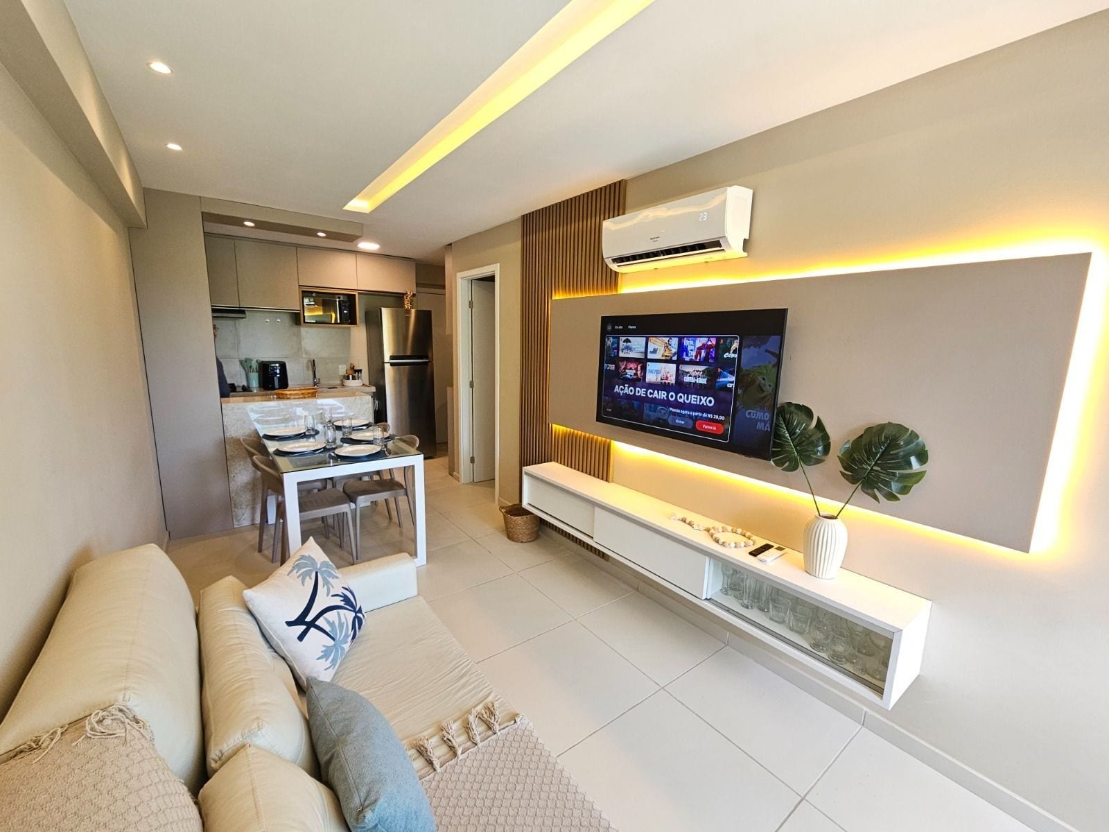 A modern living room with a flat-screen TV mounted on the wall, a sofa, and a dining area with a kitchen in the background.