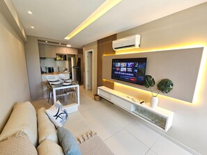 A modern living room with a flat-screen TV mounted on the wall, a sofa, and a dining area with a kitchen in the background. - Galapagos Beach Residence - Praia dos Carneiros (Tamandaré)