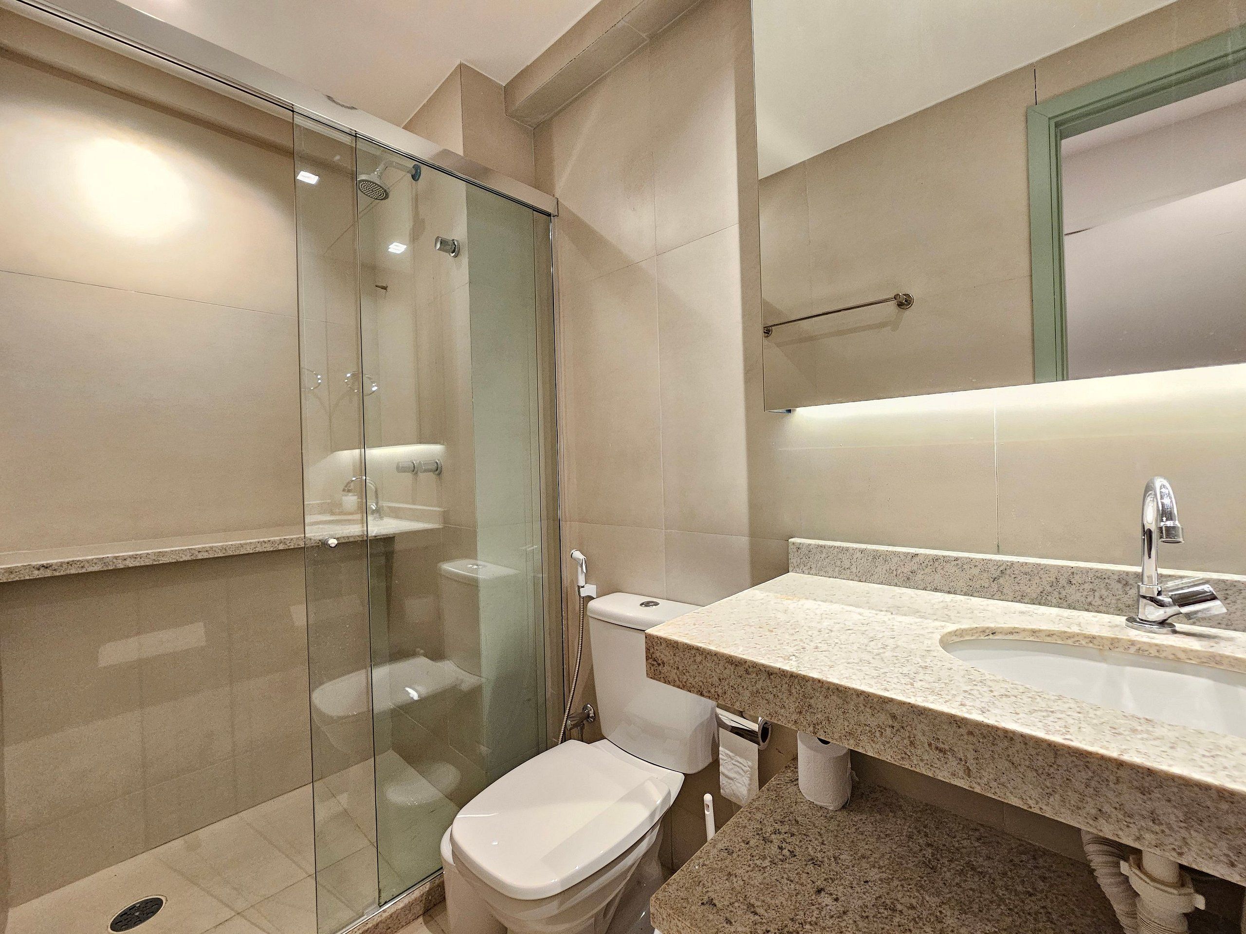 Un baño con ducha de vidrio, inodoro, lavamanos con espejo y perchero para toallas.