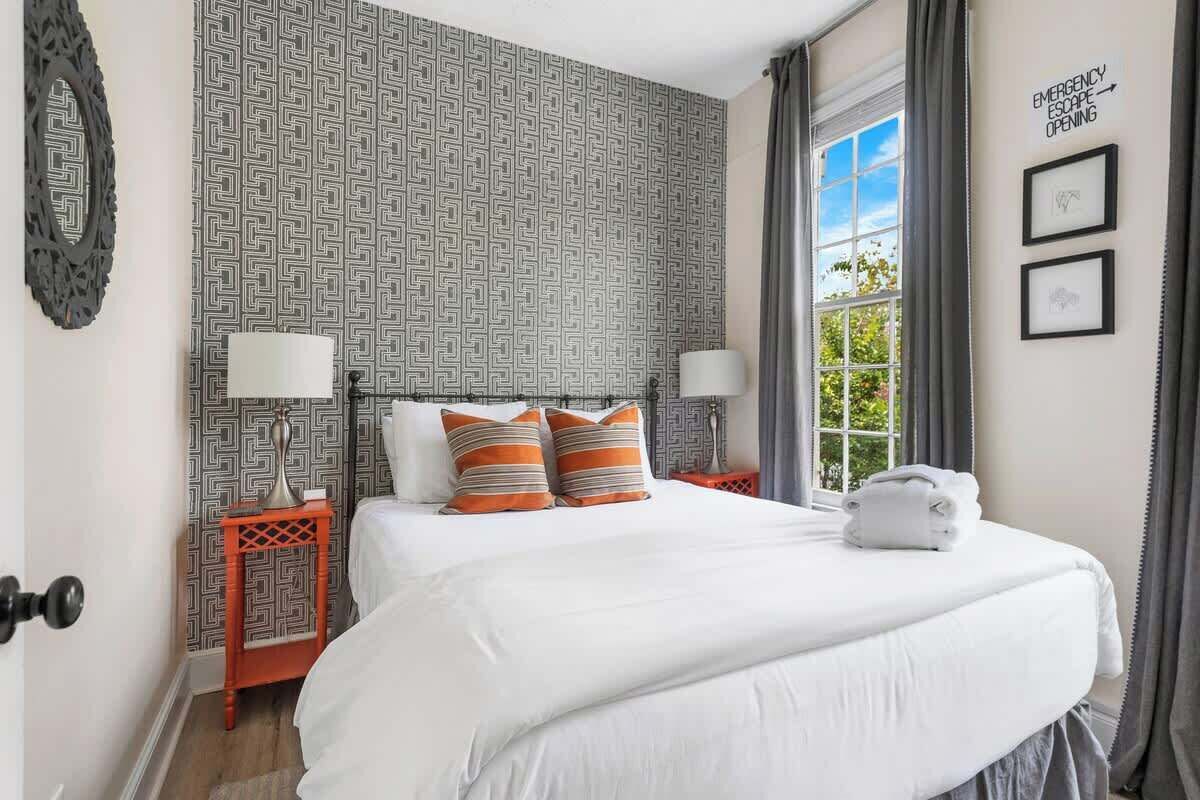 A neatly made bed with white bedding and striped pillows, a nightstand with a lamp, a window with curtains, and a wall with a geometric pattern and emergency escape sign.