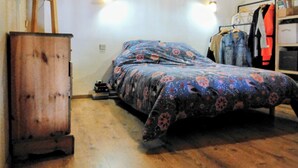 Un dormitorio con suelo de madera, cama con ropa de cama floral, armario de madera y perchero con ropa colgada.