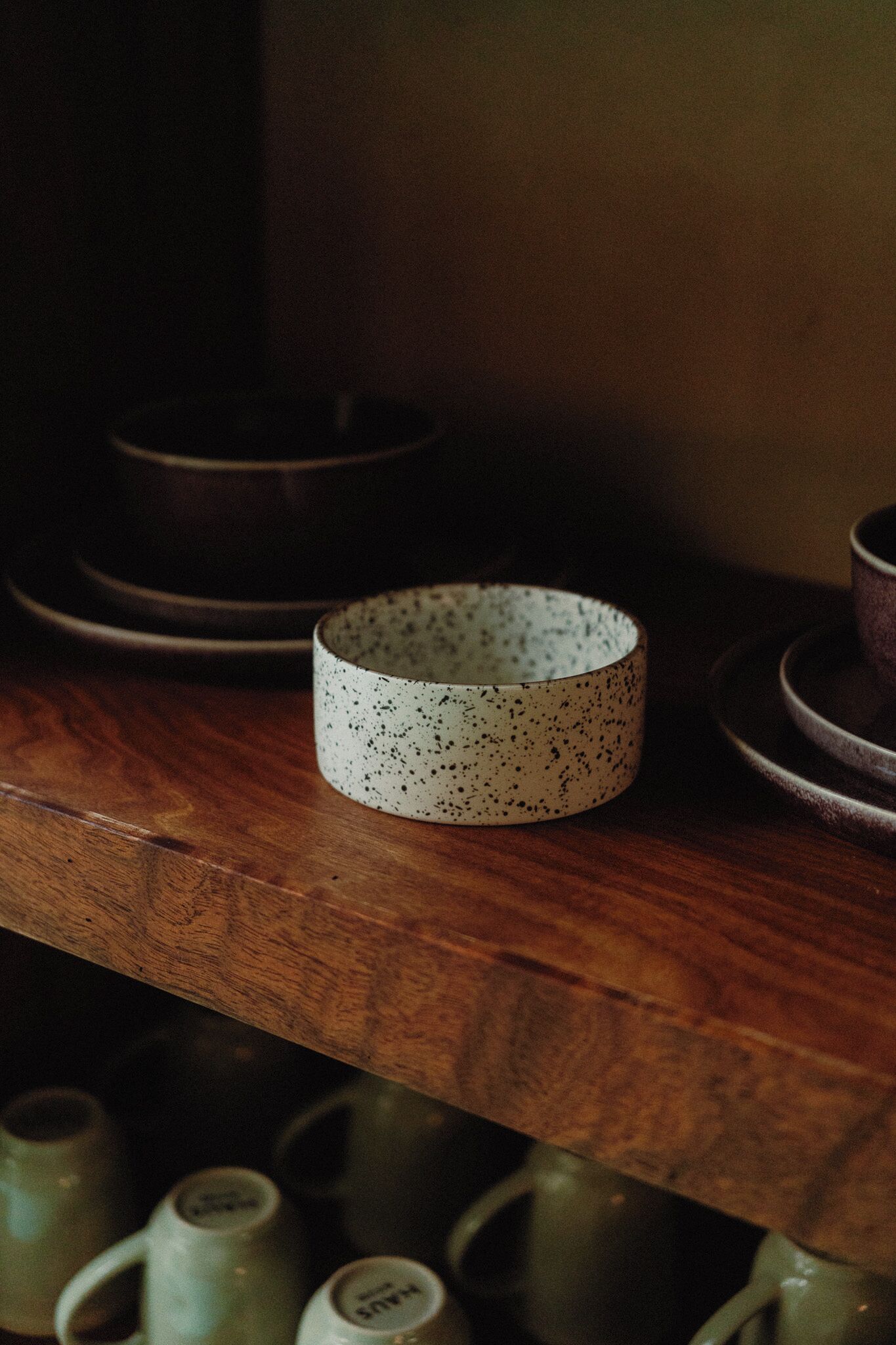 A wooden shelf with a white speckled bowl and several plain green mugs.